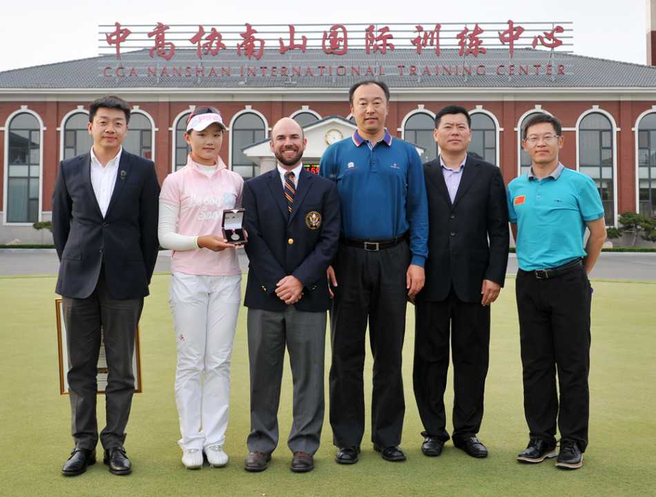 石昱婷闪耀美国女子公开赛的荣耀时刻 石昱婷面带灿烂笑容,与身着正装的颁奖嘉宾并肩而立,共同手持纪念品,身后是绿意盎然的球场与湛蓝天空,象征着荣誉与友谊的交汇。