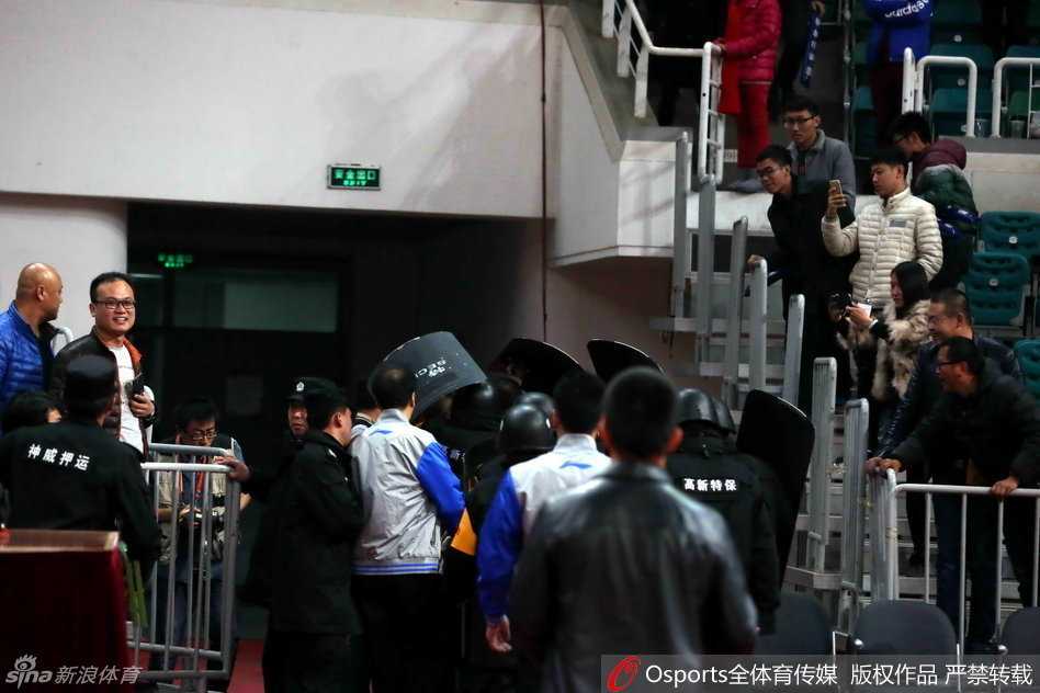 在安保人员的层层护卫下，裁判员低着头快步疾走，试图快速穿过球员通道入口，身后的背景是模糊不清但又充满压迫感的看台人群。