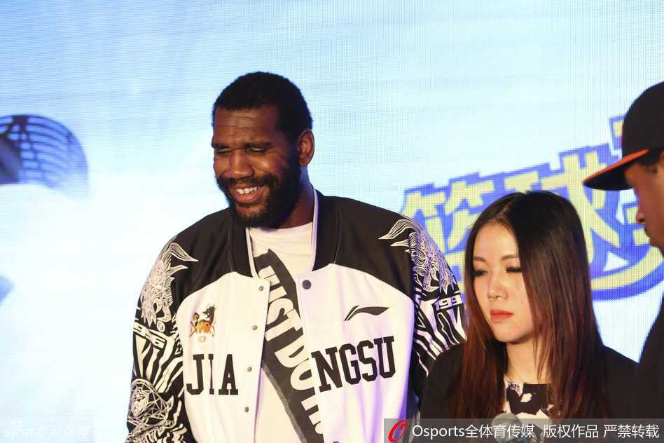 肯帝亚双星闪耀海上篮球嘉年华 胡雪峰携奥登跨界启航 奥登双手抱胸站立台上,脸上绽放出灿烂笑容,身旁的胡雪峰正在与工作人员交谈,奥登被现场气氛感染,眼中闪烁着轻松愉悦的光芒。