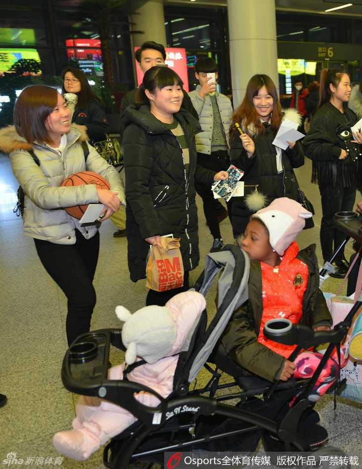 哈德森的女儿坐在婴儿车里，身穿粉色棉衣，头戴毛线帽，圆嘟嘟的脸蛋上挂着好奇的笑容，小手伸出车外，萌态可掬。