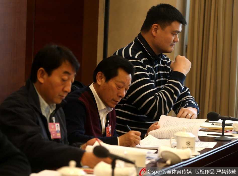 一身深色西装的姚明被记者围在人民大会堂前的空地上，他微微低头，神情专注地听着记者提问，四周的话筒和录音笔如林，几乎要贴到他身上，他身后不远处就是巍峨的建筑。