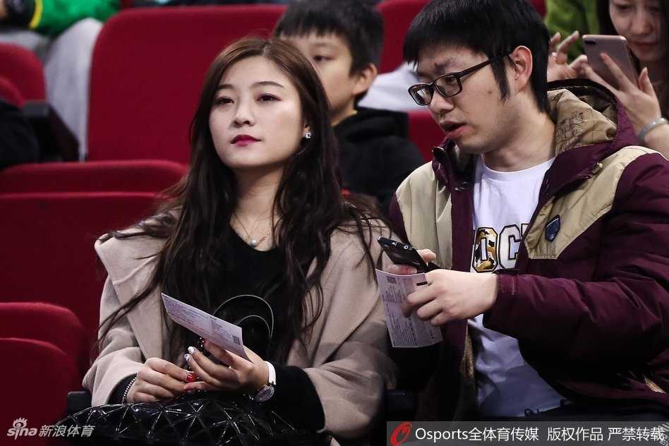 在人群的缝隙中，一位戴着黑框眼镜的美女球迷神情专注地凝视着远方。她的手里拿着北京男篮的应援围巾，安静的气质与身后喧嚣的看台形成一种独特的和谐，仿佛一位冷静的记录者。