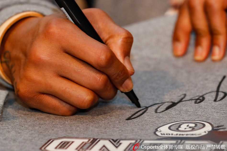 马布里为球迷签名的特写，他手中的笔正在一件卫衣上划过，脸上带着专注而满足的微笑，旁边摆放着球迷精心准备的鲜花和礼物，每一样礼物都承载着球迷沉甸甸的爱。