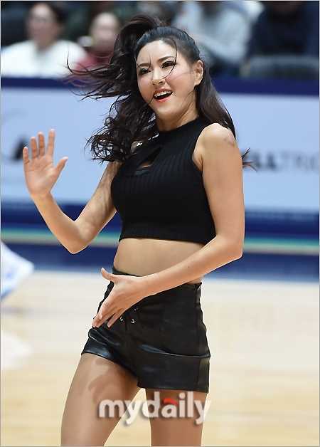 镜头拉近，捕捉到一位队员自信从容的面部表情，她手持花球，即使在高强度的舞蹈中依然保持着迷人的微笑，皮短裤的帅气与女孩的柔美在她身上完美融合