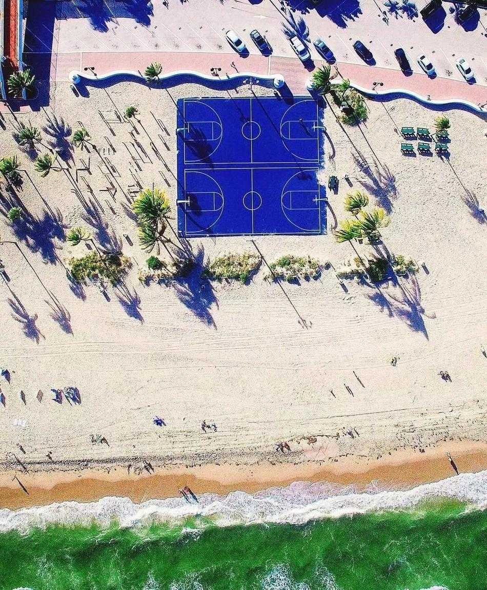 俯瞰光影交错,探寻篮球场的几何诗意 夕阳余晖下,从高处俯瞰篮球场,暖色调的灯光与蓝色地面形成鲜明对比,球员的影子被拉长,如同舞台上的舞者。