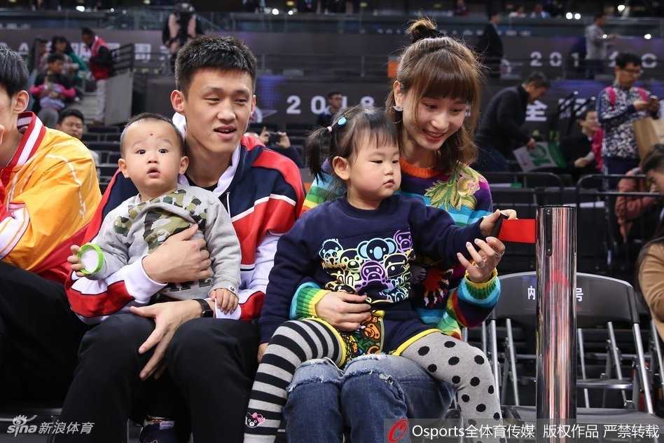 身着深色休闲装的周鹏将儿子轻抱在怀中，眼神温柔地看向镜头，身旁的女儿乖巧站立，妻子刘美则长发披肩面露微笑，一家四口在球场背景下显得格外温馨和谐