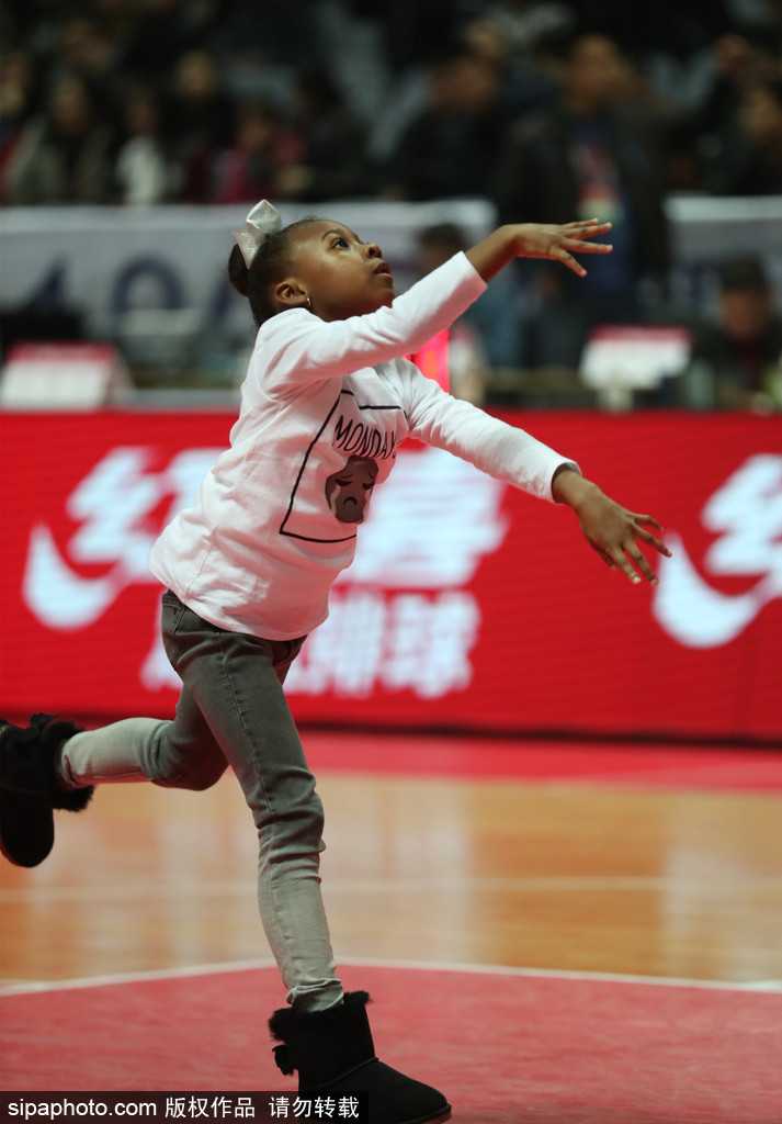 小姑娘双手抱着一个装满牛奶的粉色奶瓶，一边喝水一边目不转睛地盯着球场方向，腮帮子鼓鼓的，模样十分惹人喜爱。