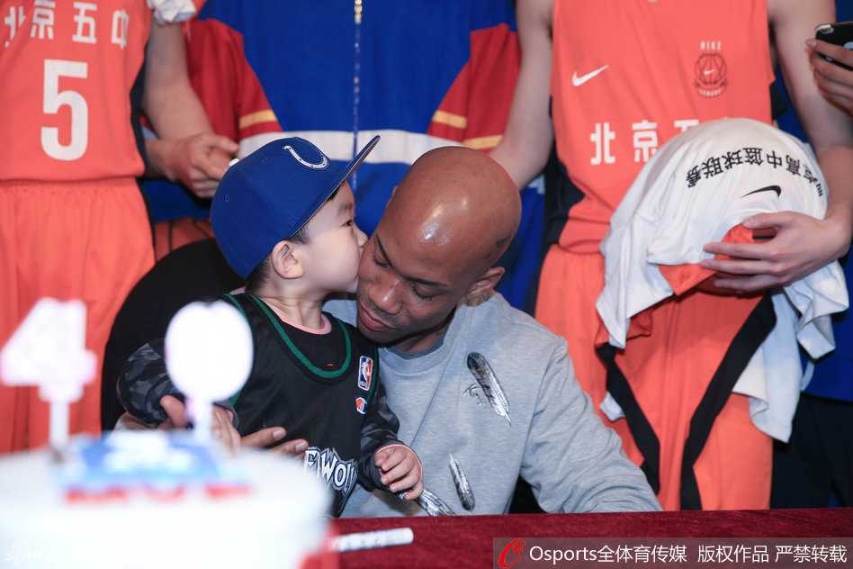 一位身穿马布里森林狼队复古球衣的小球迷被老马宠溺地抱在怀中，小家伙开心地亲吻老马的脸颊，老马则满脸享受，眼神中透出父亲般的温柔。