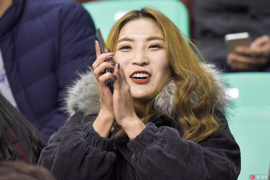 一位长发披肩的优雅女球迷，面庞在体育馆灯光的映照下显得格外立体，她目光如炬，仿佛在审视场上每一个战术变化。