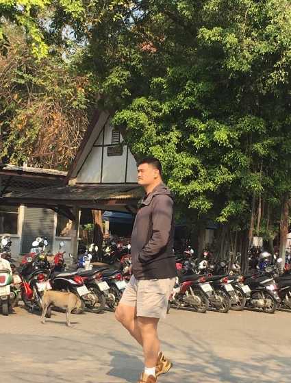 姚明独自行走在泰国清迈的街头，他身穿深色上衣配短裤，背着双肩包，高大的身影在异国建筑映衬下宛如一座移动的铁塔。周围的游客在他身后显得格外娇小，纷纷驻足侧目，完美诠释了什么叫走到哪里都是焦点。