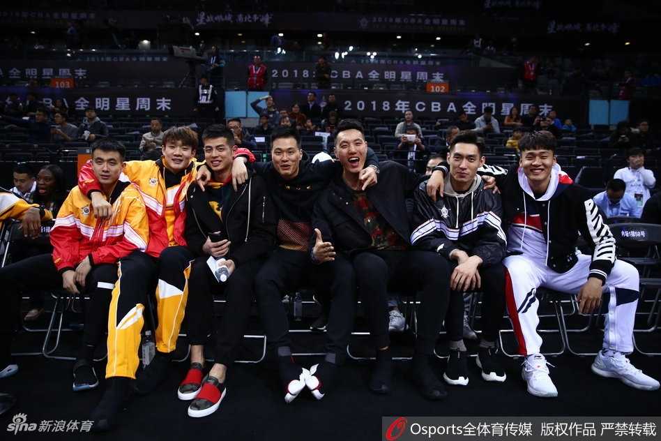 丁彦雨航与身旁的队友在观赛间隙神情放松地交谈，身后是座无虚席的深圳球迷，现场气氛热烈而轻松。