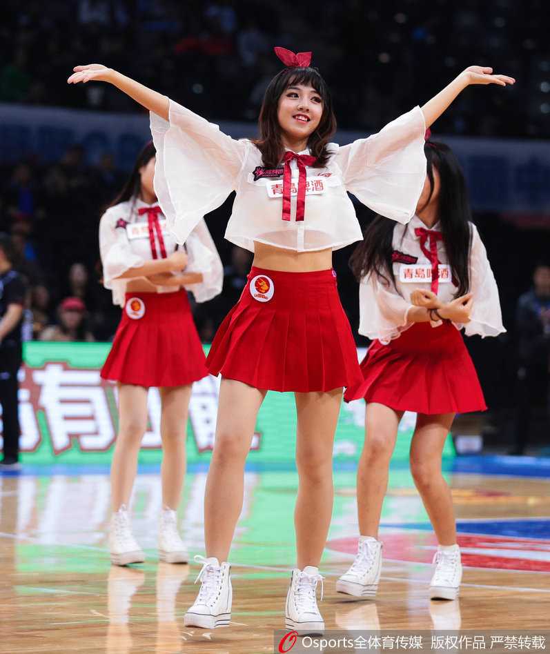 深圳篮球宝贝烈焰红裙舞动赛场 助力新世纪大胜青岛双星 表演中的篮球宝贝们动作整齐划一,她们高高跃起,红色裙摆在空中飞扬,展现出青春洋溢的动感与活力,背景是模糊的赛场看台。