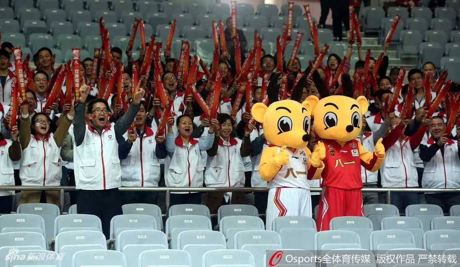 吉祥物背后抱住一位正专注于赛场的老球迷，突然的“袭击”让这位大叔先是一惊，随即绽放出灿烂的笑容，旁边坐着的朋友们也开怀大笑，看台上的氛围瞬间被引爆