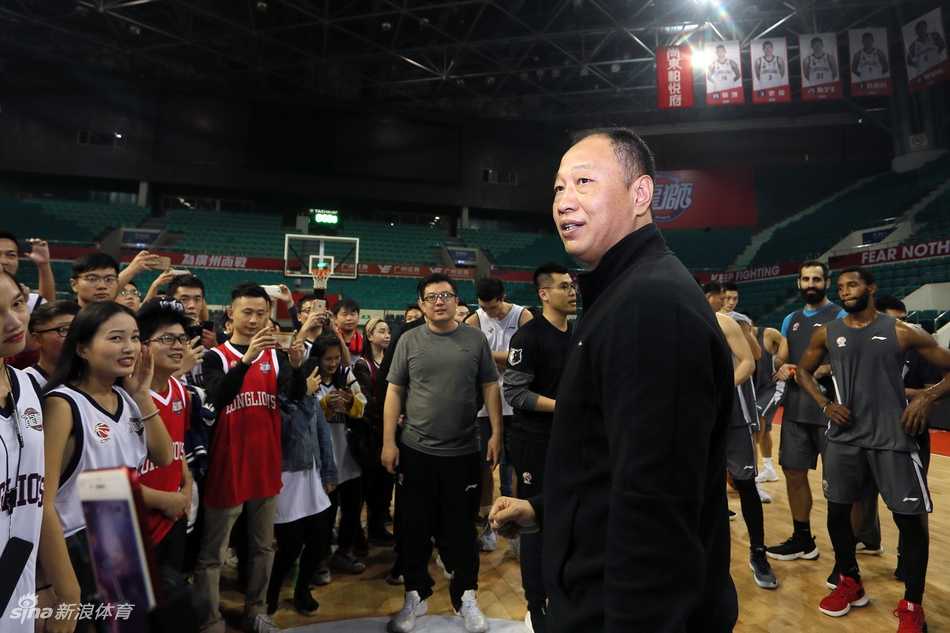 广州龙狮男篮开放日现场，球迷们围坐在场边，专注地看着场地中央正在进行的训练，背景是明亮的篮球馆和悬挂的旗帜。