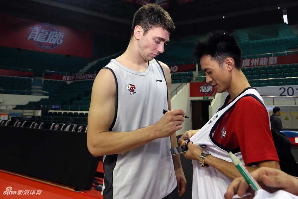 广州男篮开放日的趣味对抗环节，一名球员高高跃起试图封盖队友的投篮，旁边其他的队友和场边的球迷都被这一瞬间逗乐，气氛欢快。