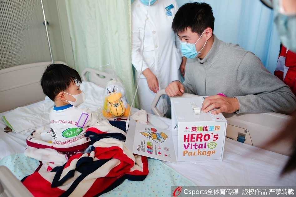 翟晓川、张松涛和吉祥物霹雳鸭一起，手捧着装满礼物的英雄能量包，准备走进病房送给孩子们，他们的脸上洋溢着温暖而真诚的微笑，背景是贴满卡通图案的医院走廊