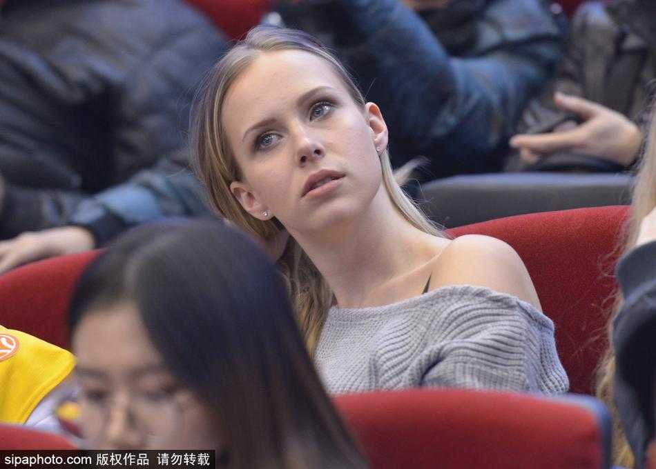 金发飘逸的莫泰妻子身着一袭深色休闲装，坐在看台上露出明媚笑容，仿佛冬日里的一抹暖阳照耀着球场