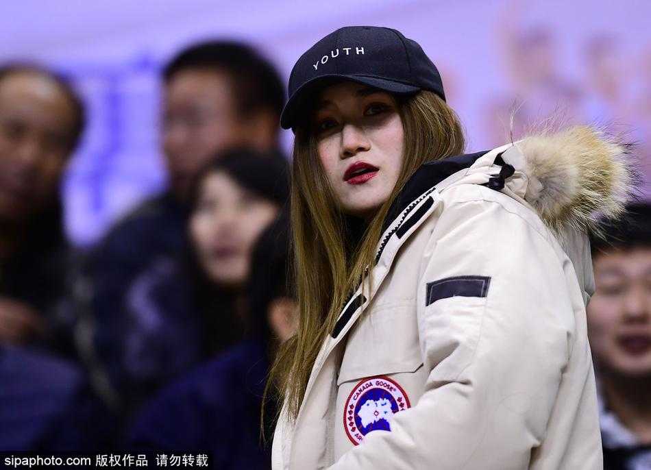 一位长发披肩的美女球迷坐在看台上，她身姿挺拔，穿着时尚的黑色外套，正全神贯注地望向赛场方向，眼神中带着对主队辽宁男篮的无限关切，仿佛下一秒就要为球员的精彩表现起身呐喊。