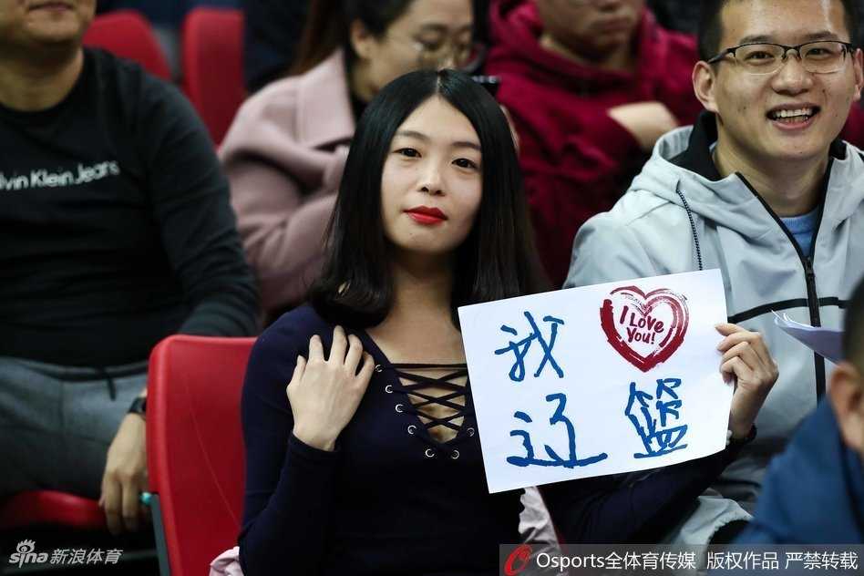 一位气质文静的辽宁美女球迷，面带微笑坐在看台上，身后是喧嚣的人群，她的笑容为紧张的比赛增添了一抹亮色。