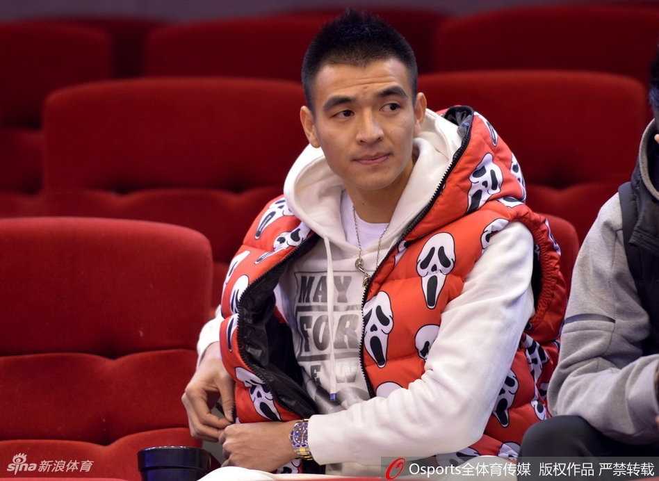 看台座位上，一位穿着时尚白色外套的年轻女球迷，手持橙色加油棒，侧身与同伴交谈，脸上洋溢着灿烂的笑容，仿佛对山东队的表现十分满意。